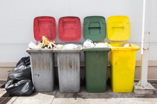 A row of four large domestic waste bins with hinged lids, positioned side by side against a white wall on a paved outdoor surface. The first bin on the left is grey and filled with miscellaneous household waste, with some rubbish spilling slightly over the top, while a black trash bag rests against its side. The second bin is also grey, appearing to contain mixed waste. The third bin is green and slightly recessed, with its lid closed, and the fourth is bright yellow, with its lid partially open, revealing some waste inside. The background shows a clean, white wall, and the bins are situated on a concrete or stone paving slab with a drain grate visible at one end. The scene subtly suggests an urban environment, typical of a commercial or residential waste collection point, common in areas like Shoreditch, London, where Commercial Waste Removal Shoreditch operates. The overall lighting indicates daytime with natural light, emphasizing the utilitarian and functional aspect of waste management in the area.