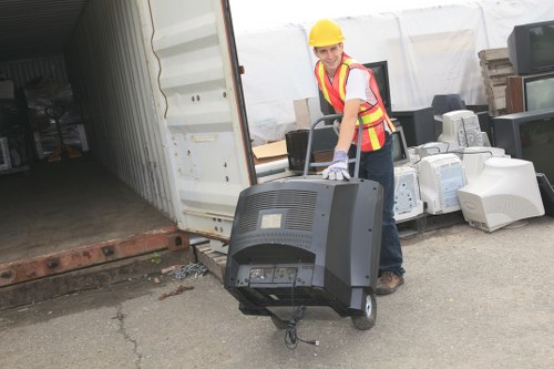 A woman wearing a yellow safety helmet, high-visibility vest, and work gloves is standing outdoors on a concrete surface near the open rear of a large shipping container. She is holding a black CRT computer monitor with an integrated stand, which is connected by a cable hanging loosely from the back. Behind her, there are multiple discarded electronic devices and appliances, including older computer towers and monitors, arranged in a pile on the ground. The background features a white temporary structure or tent, typical for waste collection or storage areas. The setting appears to be part of a rubbish removal operation, with the focus on clearing electronic waste at an industrial or commercial site, possibly in Shoreditch or nearby London.