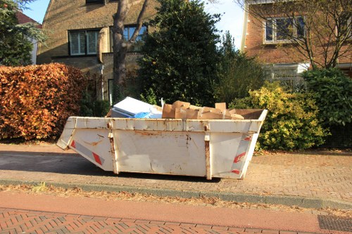A large white metal skip container filled with various flattened cardboard boxes and waste materials, placed on a brick-paved driveway along a residential street. The skip shows signs of rust and weathering, with some dirt marks on its exterior. It is positioned close to the pavement, with a backdrop of residential houses, trees, and bushes in a suburban setting. The surrounding environment includes a neatly maintained hedge on the left and a dark green shrub behind the skip, indicating a typical urban or suburban neighbourhood. The lighting suggests daytime with natural sunlight casting soft shadows, and the scene overall reflects the type of waste removal handled by Commercial Waste Removal Shoreditch in the local area of East London.