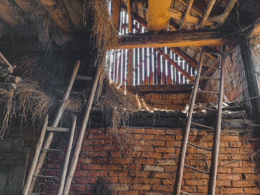 An interior view of an old, dilapidated brick structure showing a partially collapsed roof with missing wooden slats allowing sunlight to filter through. The brick walls are weathered and uneven, with